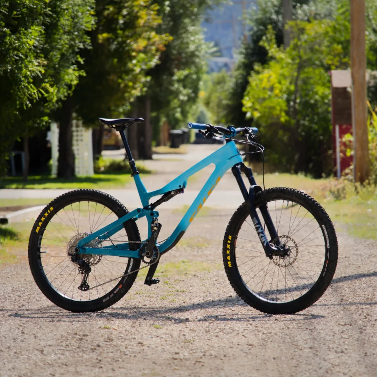 Teal 2024 Salsa Rustler full-suspension mountain bike with Maxxis tires, standing on a gravel alley lined with green trees.